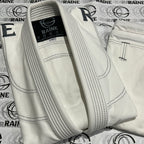 A white Brazilian jiu-jitsu gi with black stitching and grey embroidery, laid out on a surface with 'RAINE' branding visible.