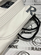 A white Brazilian jiu-jitsu gi with black stitching and grey embroidery, laid out on a surface with 'RAINE' branding visible.